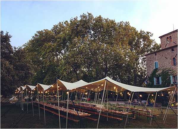 An outdoor wedding reception setup on the grounds of a French stone château, photographed at dusk with a wide establishing shot. A large stretch tent with a white undulating canopy is erected over two long wooden banquet-style trestle tables with bench seating, set and dressed for dinner service. Warm-toned string lights hang beneath the tent canopy, providing ambient lighting as the natural light fades. The rustic, informal dining style with wooden furniture suggests a relaxed, country aesthetic. Potential venue feature image.