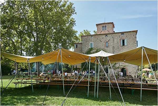 An outdoor wedding reception set up on the grounds of a historic stone tower or small chateau, photographed in a wide shot. Two large yellow stretch tent canopies cover long banquet-style tables with green metal bench seating, arranged in parallel rows on a lawn. The tables appear to be set with glassware and decorative items, and a small group of guests or staff are visible in the background near the building. The setting has a rustic, informal feel with the medieval stone architecture as a backdrop. Potential venue feature image.