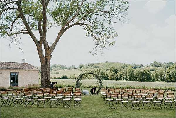 An outdoor ceremony setup being prepared on a lawn, with two people arranging a circular greenery arch at the altar point. Rows of dark metal bistro-style chairs are arranged in two sections facing the arch, with capacity for approximately 80–100 guests. A small pale stone outbuilding is visible to the left, and open countryside with rolling fields stretches across the background. The styling is relaxed and rustic, with the greenery-only circular arch as the focal ceremony structure. Wide shot. Potential venue feature image.
