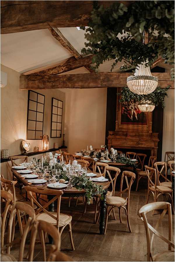 An indoor wedding reception room set for dinner, featuring two long wooden farm tables lined with cross-back wooden chairs with woven rush seats. The tables are dressed with lush greenery runners, white pillar candles, glassware, and white plate settings. A beaded chandelier hangs from the ceiling alongside a large overhead greenery installation. The room has exposed wooden ceiling beams, warm ambient lighting from candles and wall sconces, black-framed window-style mirrors on the wall, and a large ornate wooden fireplace as a focal point. The styling is rustic with warm, earthy tones throughout. Wide interior shot taken from a slightly elevated angle. Potential venue feature image.
