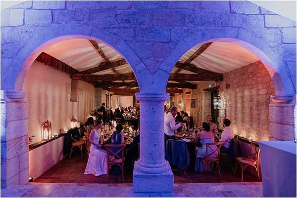 A wedding reception dinner is underway inside a historic stone venue, photographed through two large arched openings supported by a central stone column. The interior features exposed stone walls, wooden beam ceilings, and cross-back wooden chairs around tables dressed with royal blue linens. Guests are seated and dining, with the bride visible in a white dress on the left side. The space is lit with dramatic blue and pink uplighting against the stone walls, supplemented by lantern-style candles on the tables. The wide-angle shot is taken from outside the arched facade, framing the entire reception room and creating a symmetrical composition. Potential venue feature image.