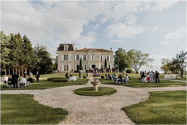 A cocktail hour or outdoor reception is taking place on the formal grounds of a classic French chateau, with approximately 50–60 guests mingling, seated, and gathered in small groups across a wide lawn. The chateau is a two-storey limestone building with a terracotta-tiled roof, tall windows, and symmetrical facade typical of Bordeaux-region architecture. The formal garden in the foreground features a gravel path, a stone fountain as a central focal point, and manicured circular lawn panels. White cocktail tables, white lounge seating, and folding chairs are arranged across the grounds. The image is a wide exterior shot taken from ground level, capturing the full scope of the venue, its grounds, and the guest activity. Potential venue feature image.