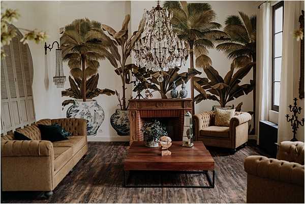Interior shot of a venue lounge or sitting room styled for a wedding event, featuring no people. The room is decorated in a colonial tropical style with a large hand-painted or wallpapered mural of palm and banana leaf trees covering the back wall. A crystal chandelier hangs from the ceiling above a wooden coffee table styled with greenery and small decorative objects. Two camel-colored tufted Chesterfield sofas and a matching armchair are arranged around the coffee table, accented with teal and gold throw pillows. A carved wooden fireplace mantel sits beneath the mural, flanked by large blue-and-white ceramic vases. The flooring is dark wood, and wrought iron candelabras and wall sconces add to the moody, vintage aesthetic. Wide interior shot. Potential venue feature image.