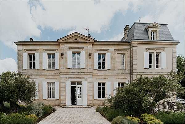 Wide exterior shot of a classic French chateau featuring cream-colored limestone façade with white shutters, a symmetrical two-story design with a central pediment, slate mansard roof with dormer windows, and a cobblestone pathway leading to the front entrance. The building displays traditional 18th-19th century French architectural details including tall French windows and decorative stonework. No wedding party or guests are visible in the frame. Potential venue feature image.