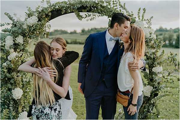 An outdoor post-ceremony moment showing the bride and groom receiving congratulations from two female guests beneath a circular floral arch. The arch is decorated with eucalyptus greenery and white flowers including hydrangeas and roses. The groom wears a navy three-piece suit with a light blue bow tie, while the bride wears a white gown. On the left, the bride embraces a guest in a dark floral dress, and on the right, the groom leans in to kiss the cheek of a guest in a white top and jeans who is carrying a tan crossbody bag. The setting is an open green field with trees visible in the background. The shot is a medium wide portrait taken at ground level, capturing all four people within the frame of the arch.