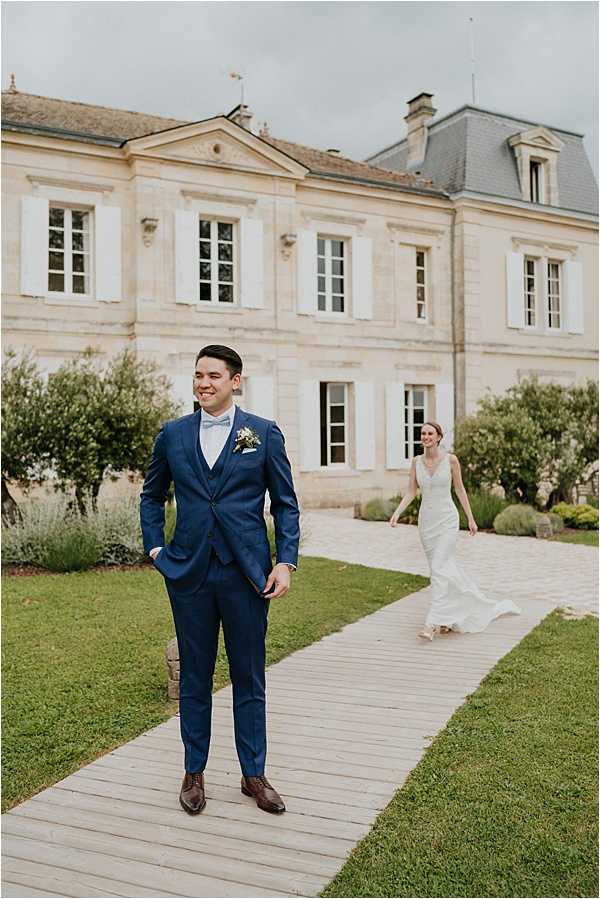 A first-look moment captured outdoors on a wooden pathway leading up to a classic French chateau with limestone facade, grey mansard roof, and white shuttered windows. The groom stands in the foreground wearing a fitted navy blue three-piece suit with brown leather oxford shoes, a light blue bow tie, and a small mixed floral boutonniere; he is smiling and looking to the side. The bride approaches from behind along the pathway, wearing a fitted ivory lace gown with a V-neckline and a short train. The composition is a full-length portrait shot with the groom sharp in focus and the bride slightly soft in the background, giving depth to the image. Potential venue feature image.