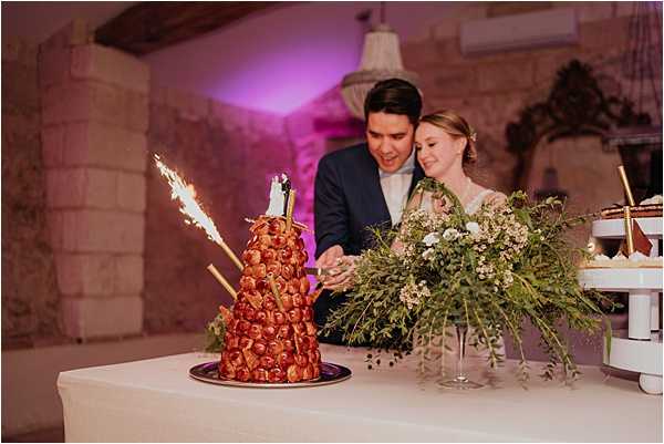 The bride and groom are cutting a traditional French croquembouche at their wedding reception, set in an indoor stone-walled venue lit with pink/purple uplighting and a chandelier overhead. The croquembouche is decorated with sparklers and topped with a small bride-and-groom figurine, displayed on a white linen-covered table alongside a loose arrangement of greenery and small white florals. The groom wears a navy suit and the bride wears a lace or embroidered gown with her hair up. The shot is a medium close-up capturing both the couple and the dessert table, with a tiered cake stand visible to the right.