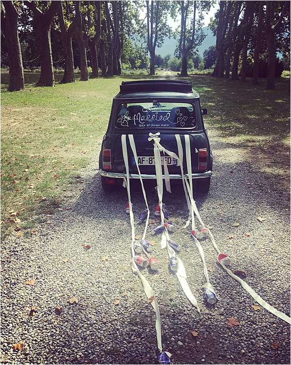 A dark green classic Mini Cooper serves as a decorated wedding getaway car, photographed from behind on a gravel driveway lined with tall trees. The rear window is painted with 'Just Married' text surrounded by hand-drawn hearts, and multiple white ribbon streamers trail from the back bumper along with several tied tin cans dragging along the gravel. The setting appears to be the grounds of a French estate or chateau, with a tree-lined avenue stretching into the distance. This is a wide exterior shot with no people visible, focusing entirely on the decorated vehicle as a wedding detail.