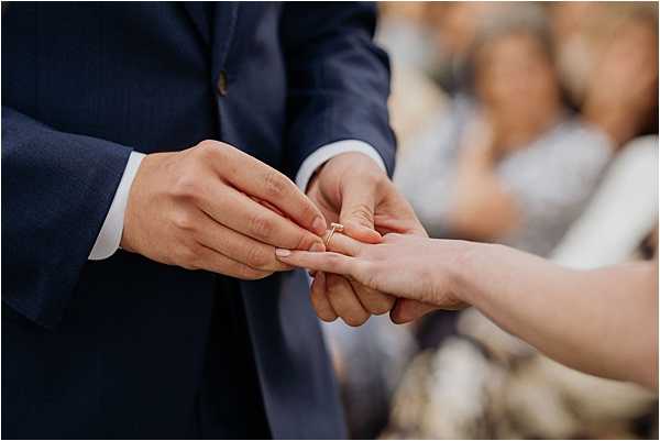 Close-up detail shot of the ring exchange moment during a wedding ceremony, showing a groom in a navy blue suit placing a thin rose gold wedding band onto the bride's finger. The bride's hand is extended toward him, and seated guests are visible in soft focus in the background. The composition isolates the hands at the center of the frame, emphasizing the rose gold ring as the focal point.