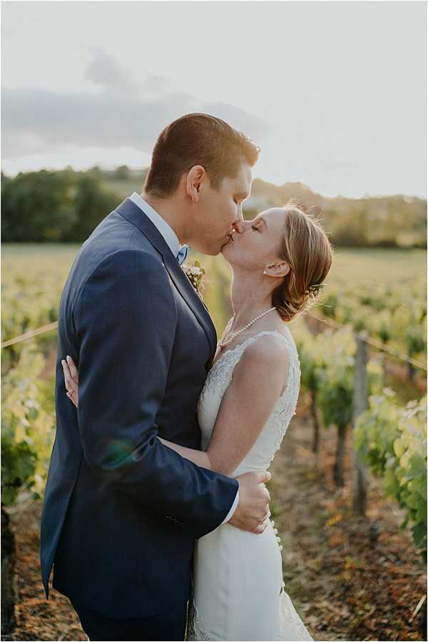 A couple portrait taken during golden hour in a vineyard, with the bride and groom sharing a kiss among the vine rows. The groom wears a navy blue suit with a small floral boutonniere, while the bride wears a fitted white lace gown with a sleeveless design and accessorizes with a pearl necklace, her short auburn hair styled with a small hair piece. The warm backlight creates a sun flare effect across the frame, softly illuminating both figures against the out-of-focus vineyard rows stretching into the background. This is a mid-distance portrait shot with a slightly shallow depth of field.