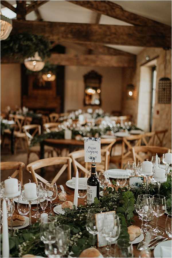 Indoor wedding reception setup in a rustic barn venue with exposed wooden ceiling beams and warm ambient lighting from pendant chandeliers. The long wooden dining tables are styled with lush greenery runners featuring eucalyptus and small white filler flowers, white pillar candles on glass candleholders, tall white taper candles, wine glasses, white plates, and a wine bottle used as a table number holder displaying a card reading 'Pessac Léognan' with watercolor floral detailing. Natural wood cross-back chairs surround the tables, and the overall decor palette is green and white with rustic, organic styling. Wide-angle shot taken from a low angle looking down the length of the foreground table toward the full room setup.