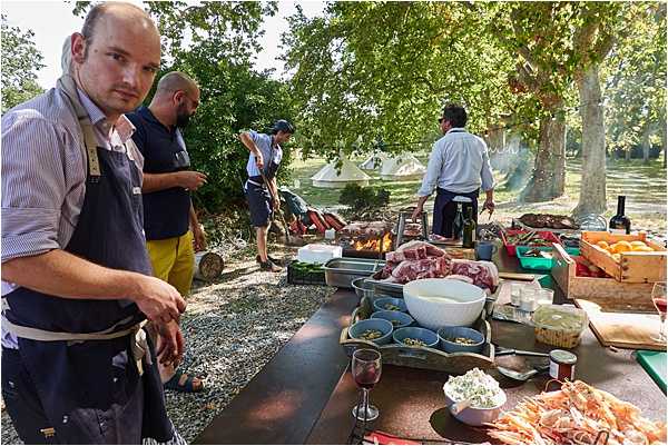 An outdoor catering or cooking setup at what appears to be a wedding reception or cocktail hour, taking place in a shaded park-like setting with large trees. Four catering staff members wearing aprons are visible, working at a long prep table and around open-fire grills in the background where smoke is rising. The foreground table is covered with raw ingredients including meat, shrimp, small bowls of garnishes, a large white mixing bowl, and wooden crates of produce, alongside a glass of red wine. The shot is a medium wide-angle image taken from a low angle near the prep table, giving a behind-the-scenes perspective on the catering operation.