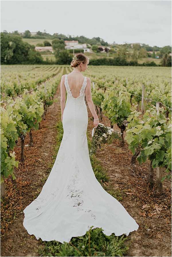 A bridal portrait taken outdoors in a vineyard, with the bride photographed from behind standing between two rows of grapevines. She wears a fitted ivory lace gown with a deep V-back, covered button detail running down the spine, and a trailing court train with lace appliqué detailing. Her hair is styled in an updo with a small floral or gold hair accessory. She holds a loose bouquet with white flowers and greenery at her side. The composition is a full-length portrait shot from behind, with the vineyard rows creating strong leading lines toward a rural landscape in the background.