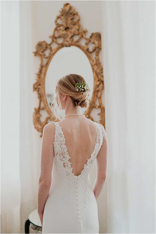 A bridal portrait taken from behind, showing the back detail of an ivory fitted wedding dress with a deep V-back neckline trimmed in lace appliqué and covered buttons running down the spine. The bride has her blonde hair styled in a low updo adorned with small white flowers, likely lily of the valley, and green foliage. She wears a pearl necklace visible at her nape. The setting is an interior room with soft white walls and sheer curtains, and the bride stands in front of a large ornate gilded oval mirror with elaborate carved gold frame. The composition is a close-to-medium portrait shot focused on the back of the dress and hair styling details.