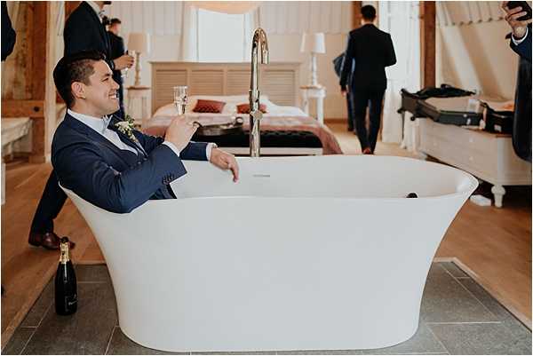 A groom dressed in a navy suit with a white boutonniere sits fully clothed inside a large freestanding white soaking bathtub, holding a glass of champagne and smiling. A champagne bottle is placed on the floor beside the tub. The setting is a well-appointed bedroom with wood flooring, white furnishings, and table lamps, where several other men in dark suits — likely groomsmen — are visible in the background getting ready, with open luggage visible. The scene captures a humorous, candid getting-ready moment. The composition is a medium wide shot taken from a low angle.