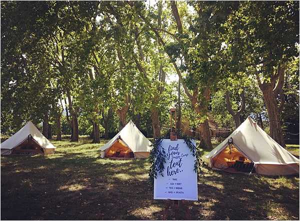 An outdoor wedding glamping setup featuring four large cream canvas bell tents arranged across a wooded area with tall mature trees. In the foreground, a white calligraphy sign on an easel reads 'find your camp tent here' with a list of guest names, decorated with greenery foliage. The tents are lit from within with warm golden lighting, suggesting an evening or late afternoon ambiance, and each tent has red/orange cushioned seating visible at the entrance. The overall styling is rustic-boho with a woodland camping aesthetic. Wide shot taken straight-on at ground level.