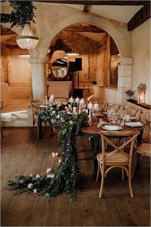 A reception table setup inside a rustic stone venue with exposed wooden ceiling beams, arched doorways, and wide-plank hardwood floors. The long dark wood dining table is styled with a lush trailing greenery garland — primarily eucalyptus — draped over the end and cascading to the floor, interspersed with small white roses and tea light candles in glass votives. White pillar candles on gold candlestick holders run the length of the table alongside wine glasses and white dinner plates. Cross-back natural wood chairs surround the table. Overhead, a beaded chandelier hangs from the ceiling, and a lantern with a lit candle is visible on a shelf in the background. The overall decor palette is dark wood, gold, and deep green with white accents, giving a rustic yet refined feel. Wide-angle interior shot. Potential venue feature image.