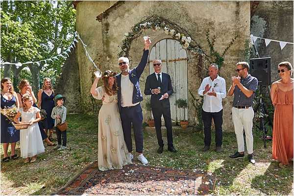 An outdoor ceremony conclusion moment in which the bride and groom raise their joined hands in celebration, standing on a vintage patterned rug in front of a rustic stone building with a weathered arched wooden door. The arch behind them is decorated with a circular floral frame of white flowers and greenery, and white triangle bunting is strung overhead. The bride wears a flowing off-white dress with floral embroidery details and a flower crown, while the groom wears navy trousers, a white shirt, and a casual navy blazer with white sneakers. An officiant in a navy suit stands just behind them, and approximately eight guests and bridal party members surround them, clapping and celebrating — including bridesmaids in navy and terracotta outfits, a young flower girl in white, and a page boy. The overall styling is relaxed boho with a natural, informal feel. Wide-angle shot capturing the full scene.
