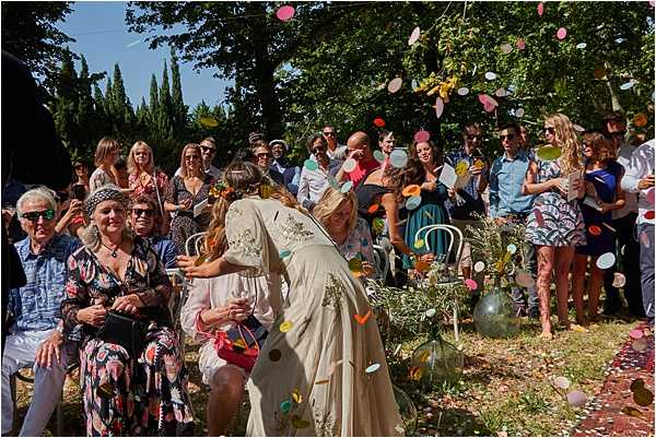 An outdoor wedding ceremony exit or aisle moment captured mid-action as guests throw large, colorful paper confetti discs in pink, orange, yellow, and green. The bride, wearing an ivory embroidered bohemian-style gown, is leaning forward to hug or interact with a young flower girl holding a small red basket. Approximately 30 guests are visible seated and standing on both sides of a grass aisle, dressed in casual summer attire in a mix of bright colors and prints, suggesting a relaxed boho-style celebration. The setting appears to be an outdoor garden or estate grounds shaded by tall trees, with teal garden chairs visible among the guests.