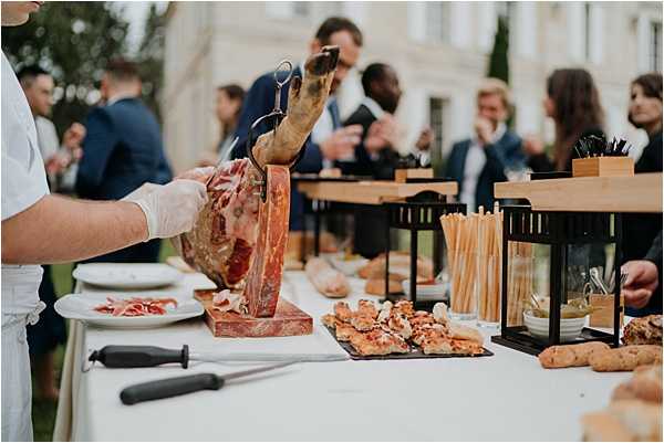 A cocktail hour food station set up outdoors, where a white-gloved server is carving a whole jamón (cured ham leg) mounted on a wooden holder, with thinly sliced portions already plated on a white dish nearby. The table is covered in a white linen and displays additional food items including breadsticks in wooden cups, black slate boards with canapés, small lantern-style condiment holders, and assorted bites. In the background, approximately 8–10 guests dressed in dark suits and cocktail attire mingle on what appears to be the grounds of a French chateau, with a white classical building visible behind them. The overall styling is classic and refined, consistent with an upscale French wedding reception. Wide shot with the food station in the foreground and guests in soft focus behind.