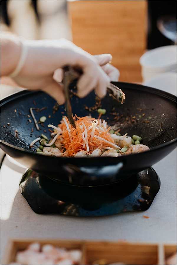 Close-up detail shot of a catering staff member wearing white latex gloves stir-frying ingredients in a black wok at what appears to be a wedding reception food station. The wok contains shredded orange carrots, white bean sprouts, edamame, and pieces of meat or seafood being tossed with tongs. The setup is placed on a black cast-iron stand over a white linen-covered surface, with raw ingredients and wooden containers visible in the blurred background, suggesting a live cooking station at an outdoor or semi-outdoor reception.