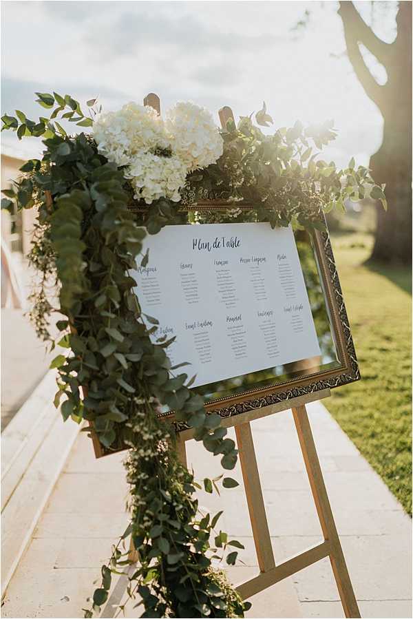 A close-up detail shot of a wedding seating chart display positioned outdoors on a paved pathway. The 'Plan de Table' is printed in calligraphy-style script on white paper and mounted in an ornate gold frame, placed on a natural wood easel. The frame is decorated with a cascading eucalyptus garland and a cluster of white hydrangeas at the top, creating a lush green and white floral arrangement. The decor palette is white and gold with abundant greenery, consistent with a classic French garden wedding aesthetic.