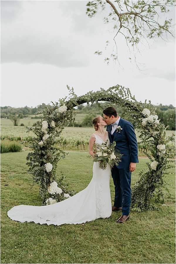 A couple shares a kiss during an outdoor portrait session in front of a circular floral arch at what appears to be a vineyard setting, with rows of vines visible in the background. The bride wears a fitted, lace wedding gown with a long train and carries a bouquet of white hydrangeas, white roses, and eucalyptus greenery. The groom is dressed in a navy blue suit with a light blue bow tie and brown leather dress shoes. The circular arch is decorated with abundant eucalyptus and mixed greenery accented by clusters of white hydrangeas, creating a green-and-white color palette consistent with the bouquet, in a classic garden style. The image is a full-length portrait shot taken at a slight distance.
