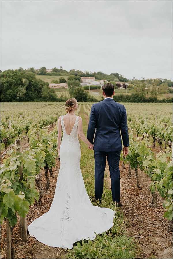 A couple portrait taken outdoors in a vineyard, with the bride and groom photographed from behind, holding hands and facing away from the camera down a row of vines. The bride wears a fitted ivory lace gown with a deep V-back and a chapel-length train, with her hair pinned up and accessorized with a small floral hair piece. The groom wears a navy blue suit. The shot is a full-length wide portrait framing both figures against rows of grapevines stretching into the distance, with a rural landscape and buildings visible on the horizon.