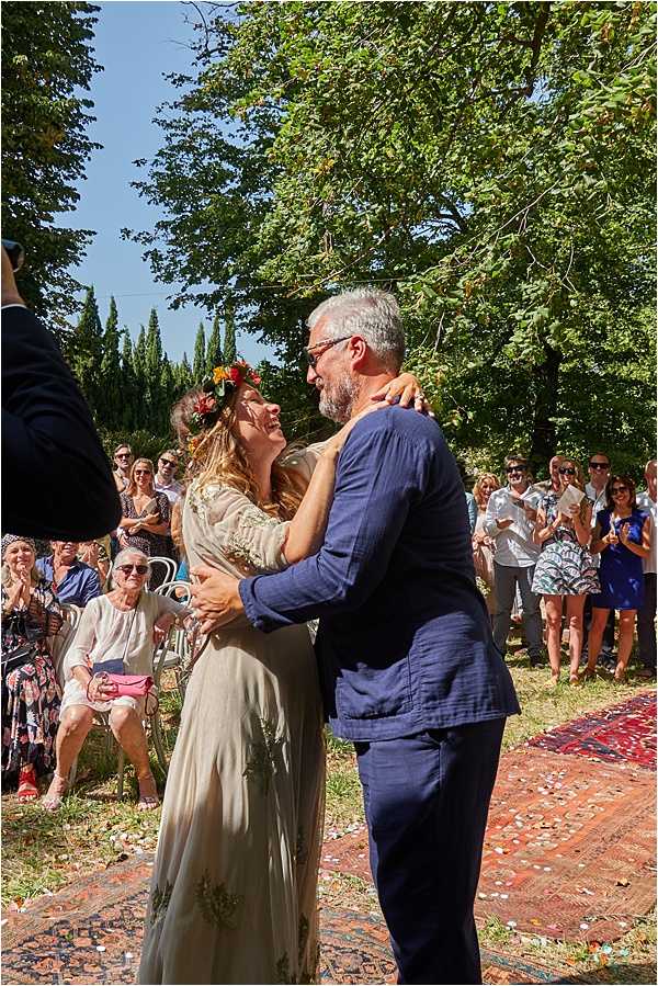 An outdoor wedding ceremony moment captured mid-portrait, showing the couple embracing and laughing together just after what appears to be their first kiss as newlyweds, with confetti scattered on the ground around them. The bride wears a loose, flowing champagne-toned dress with embroidered detailing and a bold floral crown made of red, yellow, and orange flowers, consistent with a boho styling theme. The groom wears a navy blue linen suit with no tie. Guests — approximately 25-30 visible — are seated and standing on either side of a aisle runner made from layered vintage-style red and burgundy patterned rugs, applauding and smiling, set within a tree-lined outdoor garden space.