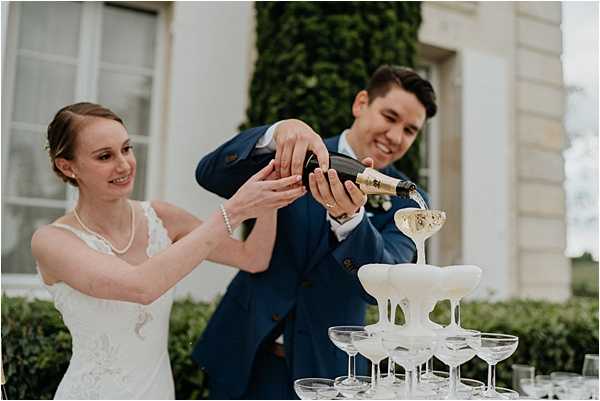The bride and groom are pouring champagne together into a coupe glass tower during an outdoor reception, with champagne visibly overflowing down the pyramid of glasses. The bride wears a white lace dress with a pearl necklace and has her hair pulled back, while the groom wears a navy blue suit with a white boutonnière. Both are smiling as they hold the bottle together. The setting appears to be the grounds of a French chateau or manor house, visible in the soft-focus background. The shot is a medium close-up portrait capturing the couple from the waist up alongside the champagne tower.