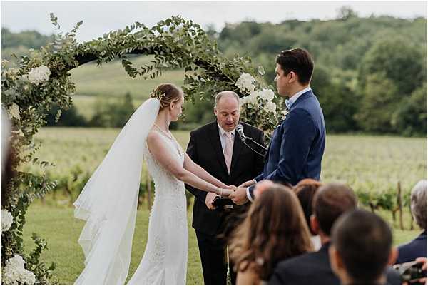 An outdoor wedding ceremony taking place in front of a vineyard, with the couple exchanging rings or vows beneath a circular arch decorated with eucalyptus greenery and white flowers, likely white hydrangeas and roses. The bride wears a fitted white lace gown with a long cathedral veil and has floral hair accessories, while the groom wears a navy suit with a light pink tie. An officiant in a dark suit stands between them, looking down, with a microphone on a stand nearby. Several guests are visible from behind in the foreground, watching the ceremony. The shot is a medium wide portrait taken from slightly behind and to the side of the guests, with the vineyard rows visible in the background.