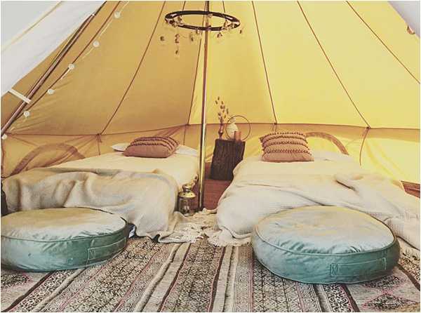 Interior detail shot of a boho-style glamping bell tent set up as wedding guest accommodation or a bridal suite. The yellow canvas tent features two low floor beds dressed in cream and white bedding with dusty mauve and taupe woven cushions, flanked by two round teal floor cushions at the foot of each bed. A rustic tree stump serves as a bedside table between the beds, holding a brass lantern and a small dried floral arrangement. A hoop chandelier decorated with hanging crystals or charms hangs from the central tent pole, and string fairy lights run along the tent seam. Layered patterned rugs in muted earth tones cover the floor, reinforcing the boho aesthetic. The overall palette is warm yellow, cream, teal, and dusty rose.