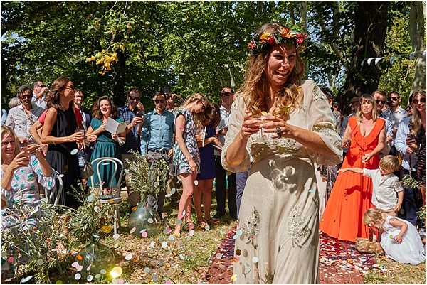 An outdoor wedding ceremony is taking place in a tree-lined setting, with the bride walking through a confetti-strewn aisle while guests line both sides cheering and clapping. The bride wears a loose, cream-colored embroidered boho-style dress and a floral crown made of orange, red, and yellow flowers in her loose wavy hair, and she appears to be laughing joyfully while holding a small glass or cup. Colorful paper confetti in pastel and mixed tones covers the ground and aisle runner. Approximately 30 guests are visible in casual to smart-casual attire, including a young child in white near the right side holding a confetti basket, and olive branches and greenery are arranged along the aisle as decor. The image is a medium-wide shot taken at ground level in bright natural daylight, with a boho-rustic outdoor styling theme.