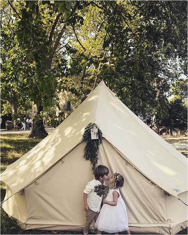 Two young children — a boy in a white shirt and tan trousers and a girl in a white tulle dress with a floral crown — share a kiss in front of a large cream canvas bell tent at an outdoor wedding. The tent entrance is decorated with a hanging garland of dark green foliage, and the girl holds a small bouquet of muted greenery and dusty blooms. The setting is a garden or estate grounds with mature trees casting dappled light, and wedding guests can be seen gathered in the background. The image is a medium portrait shot with a rustic, boho-inspired styling aesthetic.