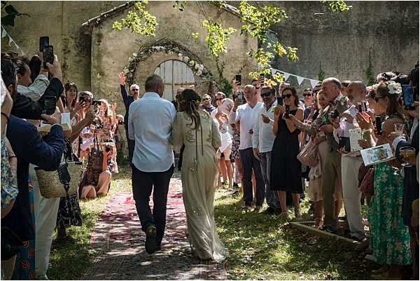 The bride and groom walk back down the aisle together following an outdoor ceremony, surrounded by approximately 30-40 guests lining both sides and cheering, many photographing the moment on phones. The ceremony takes place in a rustic outdoor courtyard in front of a weathered stone wall with a large arched doorway decorated with a floral arch of white and pale pink flowers. Colorful bunting triangular flags are strung in the background. The bride wears a long ivory or champagne-toned dress with embellished detailing and has a floral hair accessory, while the groom wears a light blue shirt and dark trousers. A patterned red and pink runner lines the aisle. The setting has a boho-rustic feel with an informal, relaxed atmosphere. Wide shot taken from behind the couple looking toward the floral arch.