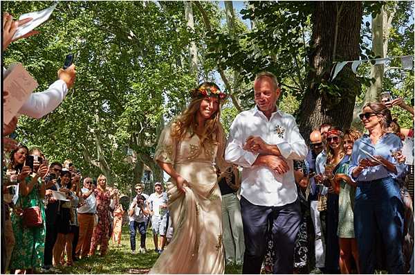 The bride and groom walk back down the aisle together in an outdoor ceremony exit under a canopy of large plane trees, surrounded by approximately 30-40 cheering and applauding guests. The bride wears a flowing champagne/gold boho-style gown with short sleeves and a colorful floral crown of orange, red, and yellow blooms; the groom is dressed casually in a white linen shirt with a floral boutonniere and navy trousers. Guests line both sides of the natural grass aisle, many holding phones to photograph the moment, and white bunting flags are visible strung between trees in the background. The shot is a wide, candid medium-distance image capturing the lively, informal boho outdoor ceremony atmosphere on a sunny day.