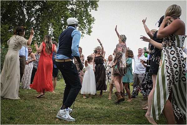 A large group of approximately 20-25 wedding guests are dancing outdoors on a grass lawn, captured in a wide, dynamic shot. The setting appears to be a garden or estate grounds, with trees visible in the background. Guests are dressed in casual summer attire including a red maxi dress, a black strapless dress, floral and patterned dresses, and a young flower girl in a white dress; one man wears a navy vest with a light blue shirt and a grey flat cap with white sneakers. The atmosphere is lively and informal, with arms raised and guests in mid-movement, suggesting an outdoor reception dance moment in a relaxed, bohemian style.