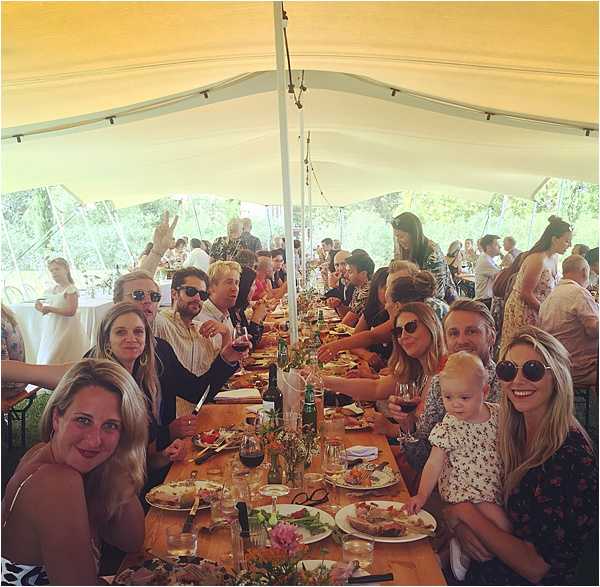 Wedding reception dinner scene photographed from the end of a long communal wooden farm table, showing approximately 30-40 guests seated and dining under a white open-sided marquee tent. The table is styled with small loose wildflower arrangements in pinks and greens, wine glasses, red wine bottles, and casual place settings on bare wood — suggesting a relaxed, rustic outdoor reception aesthetic. In the foreground, several guests including a woman holding a toddler in a white floral outfit are smiling toward the camera; a figure in white, likely the bride, is visible in the background left. String lights run along the tent ceiling, and the surrounding greenery is visible through the open sides of the marquee. Wide-angle, slightly fish-eye social shot taken from a low vantage point at table level.