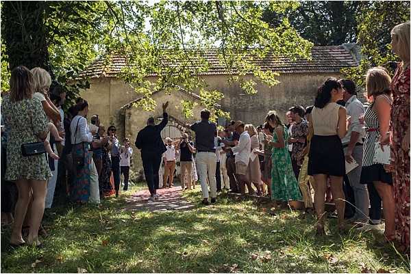 A large group of approximately 30-40 wedding guests gathers outdoors in a shaded garden area, forming two loose lines along a brick pathway, suggesting a traditional tunnel or guard of honor moment. The setting is a rustic French property with an old stone outbuilding with terracotta roof tiles visible in the background. Guests are dressed in summer wedding attire including floral dresses, fitted midi dresses, and light-colored suits and trousers. One man in a dark suit is seen with his arm raised mid-celebration. The scene is lit by dappled natural sunlight filtering through mature trees overhead. Wide-angle shot capturing the full gathering and surrounding grounds.