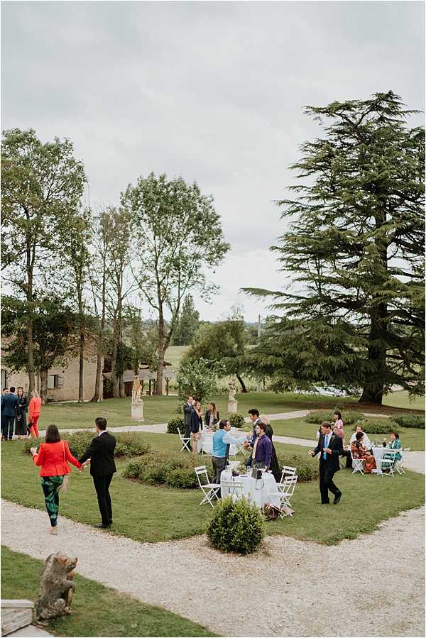 An outdoor cocktail hour taking place in a formal garden at what appears to be a French country estate or chateau grounds. Approximately 20–25 guests are mingling and seated around white folding tables and chairs arranged on a manicured lawn with low box hedge borders and stone statuary pedestals. Guests are dressed in a colorful mix of outfits including a red jacket with green trousers, a coral/orange jumpsuit, a purple dress, and various suits and ties. The shot is taken from an elevated wide angle, likely from steps or a terrace above, capturing the full layout of the garden space with a stone outbuilding visible to the left and open countryside in the background. Potential venue feature image.