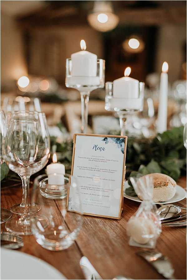 Close-up detail shot of a wedding reception table setting on a bare wooden surface. The centerpiece features a lush greenery garland running along the table, accented by multiple white pillar candles on tall clear glass candlestick holders and small votive candles in glass holders. A gold-framed French menu card with navy and dark botanical watercolor accents stands upright as a focal point. Crystal wine glasses, white side plates, silver cutlery, and a small wrapped favor with a bread roll are visible at the place setting. The overall decor palette is white, gold, and green with a classic yet natural styling approach, and the warm candlelight creates a soft, amber-toned atmosphere in the blurred background where additional table settings are visible.