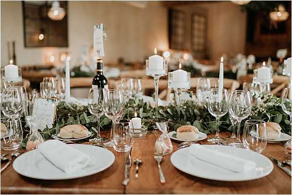 A close-up detail shot of a wedding reception tablescape set on a bare wooden farm table indoors. The centerpiece features a lush trailing eucalyptus garland running the length of the table, interspersed with white pillar candles on tall glass candleholders and white taper candles. Place settings include large white charger plates with white linen napkins, multiple wine glasses, and silver cutlery, with bread rolls already served. A bottle of red wine is visible among the decor. The overall palette is natural green and white with warm candlelight, giving a rustic yet refined aesthetic. The reception room behind is warmly lit with additional candlelight and pendant lighting visible in the soft background.