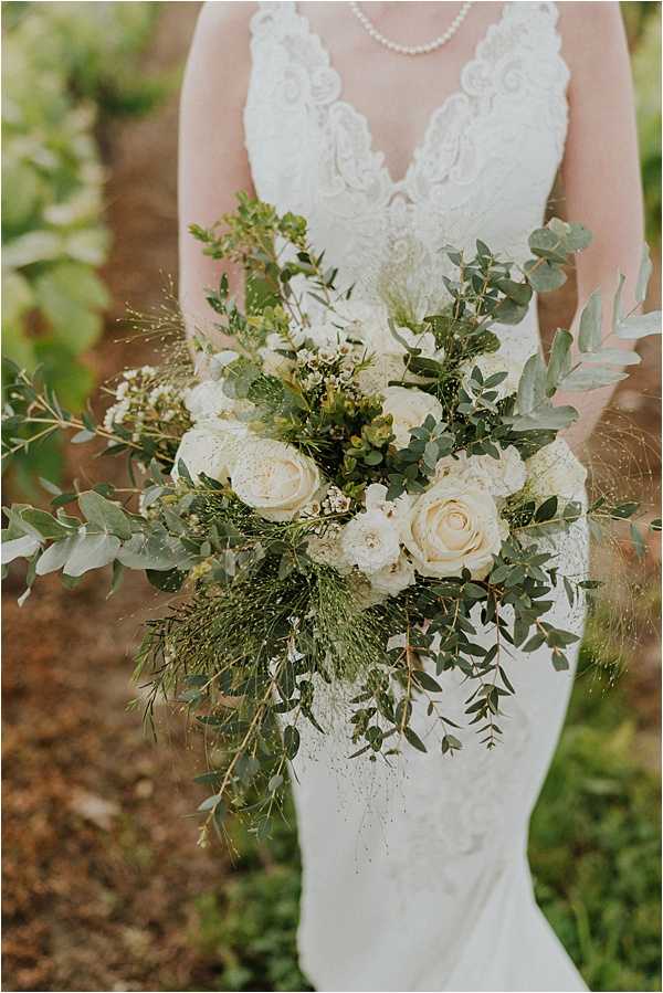 Close-up portrait of a bride holding her bouquet outdoors, with the frame cropped from the neck to the hips. She wears a fitted white lace gown with a deep V-neckline and a pearl necklace. The bouquet is a large, loosely structured cascade-style arrangement featuring cream garden roses, white ranunculus, small ivory wax flowers, eucalyptus, ruscus, and wispy feathery greenery, creating a green-and-white botanical palette with a boho-organic style. The overall styling is natural and garden-inspired.
