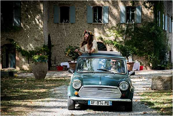 A bride and groom arrive or depart in a vintage teal/dark green classic Mini Cooper with a French license plate, photographed on a gravel driveway in front of a large stone Provençal mas or farmhouse with blue-grey shutters. The bride is standing up through the open sunroof, smiling broadly, wearing what appears to be a lace or cream-toned dress and a floral crown with dark flowers and greenery in her hair, holding a bouquet. The groom is seated at the wheel wearing sunglasses. The styling is relaxed and boho, with the vintage car as a deliberate decor choice. The shot is a medium wide portrait-orientation image taken head-on as the car approaches the camera. Potential venue feature image.