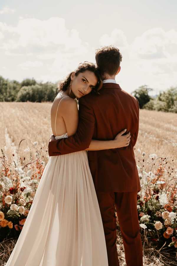 An outdoor couple portrait taken in an open field, with the bride resting her head against the groom's back and looking toward the camera while embracing him from behind. The bride wears a blush/champagne spaghetti-strap gown with a flowing skirt and a delicate floral wrist accessory, while the groom wears a deep burgundy suit with a white dress shirt. A loose floral arrangement at ground level features burnt orange, peach, cream, and deep red blooms — including what appear to be carnations, ranunculus, and wildflowers — that complement the warm tonal palette of the couple's attire. The composition is a medium portrait shot with a golden wheat field stretching into the background, giving the image a warm, boho-rustic aesthetic.