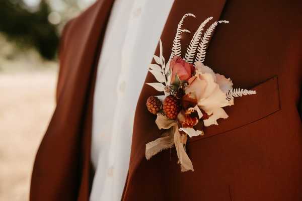 Close-up detail shot of a groom's boutonniere pinned to the lapel pocket of a rust/terracotta brown suit jacket worn over a white textured dress shirt. The boutonniere features a blush pink rose, dried blackberries or raspberry-like dark red berries, white dried fern fronds, a dried cream orchid or ranunculus bloom, and small dried foliage in warm tan and brown tones, creating an autumnal, boho-rustic aesthetic. The color palette of the floral arrangement — blush, rust, burgundy, and ivory — coordinates directly with the terracotta suit. The background is softly blurred with warm, golden outdoor tones.