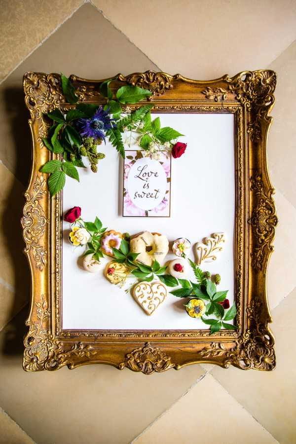 A flat-lay detail shot of a large ornate gold baroque-style picture frame laid on a tiled floor, styled as a dessert and decor display. Inside the frame, a small printed card reading 'Love is Sweet' in calligraphy script is propped at the top center, decorated with a floral border. Arranged within the frame are iced sugar cookies in heart and floral shapes with gold and white royal icing details, mini ring-shaped pastries, and small iced biscuits with red berry accents. Fresh greenery including fern fronds, raspberry leaves, and small branches with red roses, blue thistle, white flowers, and yellow blooms are scattered across the frame and spilling over its edges. The styling is romantic and garden-inspired with a rich jewel-toned floral palette against a white background insert.