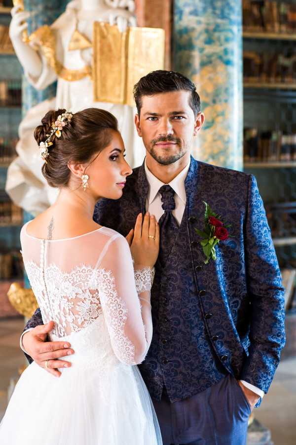 A couple portrait taken indoors in what appears to be an ornate, art-filled venue featuring a large white and gold statue and teal-painted decorative columns with bookshelves visible in the background. The bride wears a white gown with long sheer lace sleeves, a lace bodice with button-back detail, and an upswept hairstyle adorned with a floral gold and blush hair piece and drop earrings; a tattoo is visible on her upper arm. The groom wears a navy blue paisley-patterned three-piece suit with a matching tie and a deep red rose boutonniere with green foliage. The composition is a close-up portrait with the bride facing away from camera and the groom looking directly into the lens, with a classic, editorial styling aesthetic.