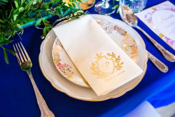 Close-up detail shot of a wedding reception place setting laid on a royal blue linen tablecloth. The setting features a gold-rimmed charger plate with a floral motif topped with a smaller matching plate, and an ivory folded napkin embossed with a gold monogram crest bearing the letter 'P' and the couple's names 'Ella & Adam.' Silver cutlery is arranged on either side, and a partially visible watercolor menu card sits at the top right corner. The overall decor palette combines royal blue, gold, and ivory, suggesting a classic formal styling with personalized stationery details.