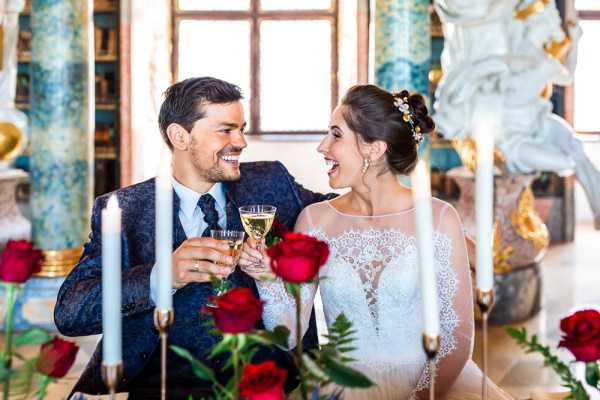 A bride and groom share a champagne toast indoors, laughing and looking at each other in a close portrait-style shot. The groom wears a navy blue patterned suit jacket with a dark tie, and the bride wears a lace illusion-neckline wedding gown with her dark hair pinned up and adorned with a small gold floral hair accessory. The foreground features tall white pillar candles in gold holders and deep red roses, creating a rich, jewel-toned decor palette. The background reveals ornate interior architecture with blue and gold decorative columns and white sculptural details, suggesting a historic chateau or palace setting.